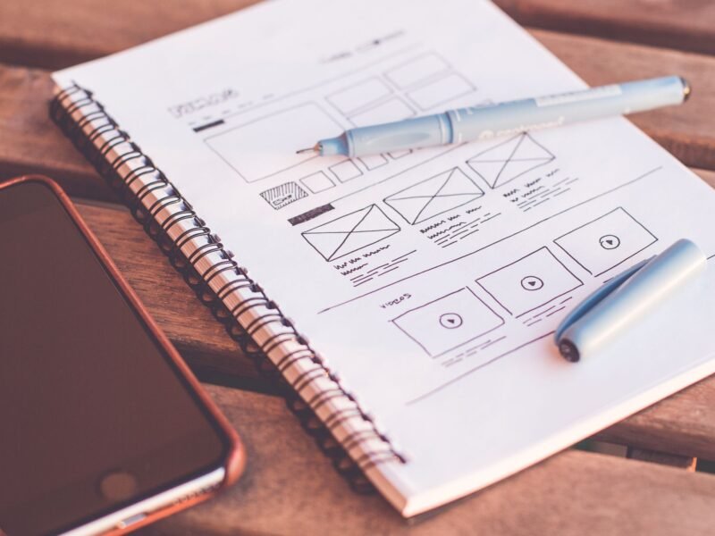 A hand-drawn website wireframe sketched in a notebook, with a pen and a phone on the desk beside it