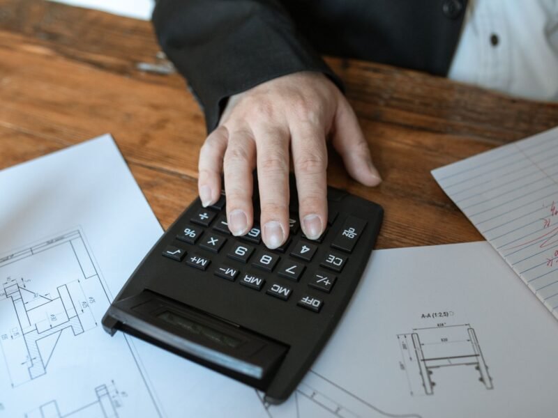 A close-up of a person working out a figure on a calculator, with technical drawings spread across the desk