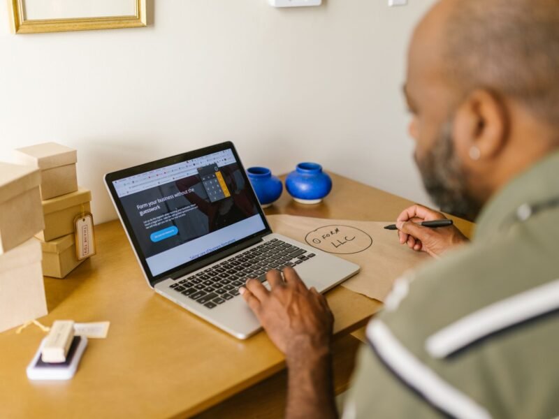 A small business owner sitting at a laptop reading a company website, with packed shipping boxes on the desk beside him
